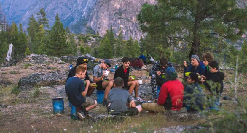 A group of students sit in a circle in a wooded area. They each have a bowl of food. 